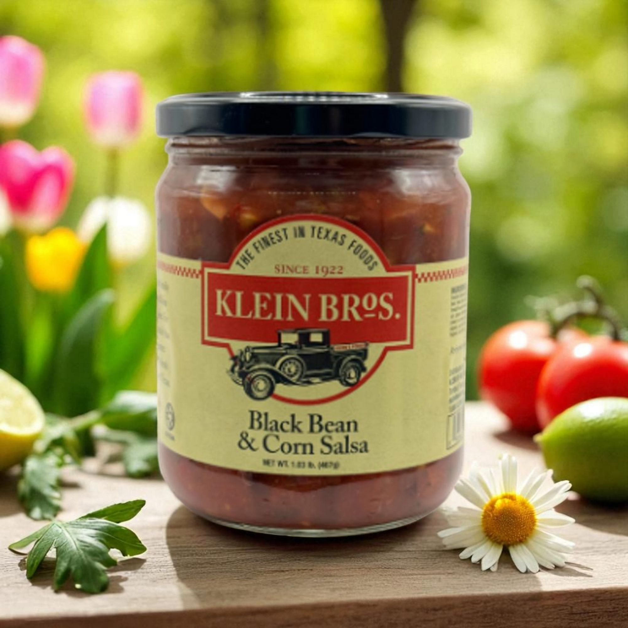 Jar of Klein Bros. Black Bean & Corn Salsa on a wooden surface with flowers and tomatoes in the background