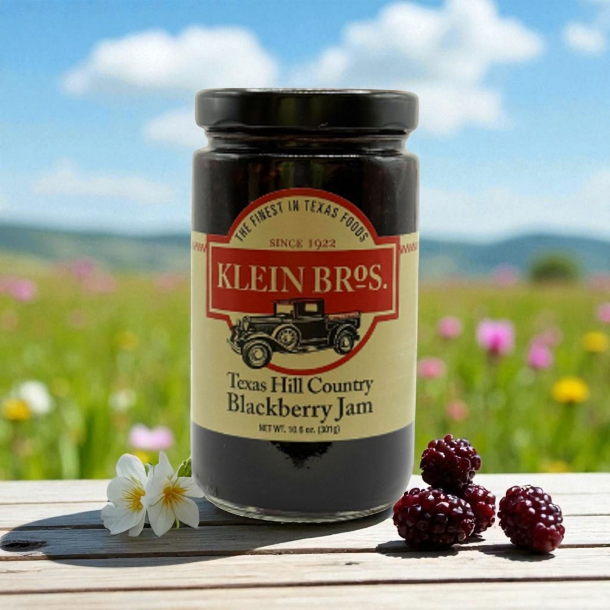 Jar of Klein Bros. Texas Hill Country Blackberry Jam with blackberries and flowers on a wooden surface with a field background.
